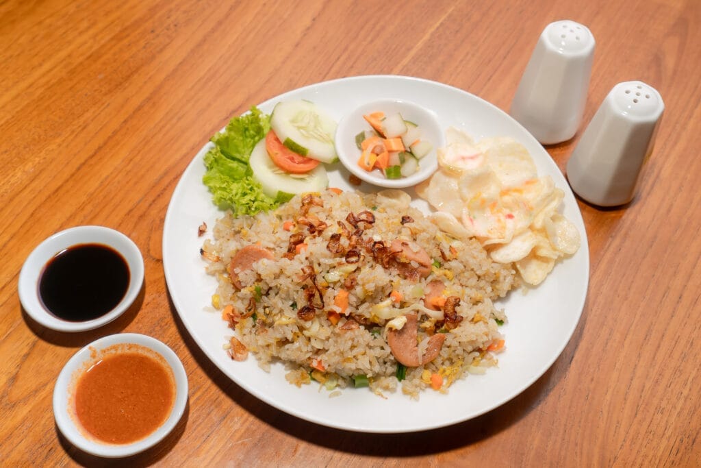 A plate of Indonesian fried rice (Nasi Goreng) with sliced sausage, crispy fried shallots, crackers, fresh vegetables, and a side of pickles, served with soy sauce and spicy sambal on a wooden table.